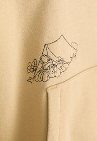 Figura di cartone con un cappello invernale che sbircia da una tenda, circondata da fiori, ricamata in nero su tessuto beige vicino a una tasca.