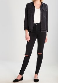 Giacca di jeans nera sopra una maglietta bianca, abbinata a jeans skinny neri con strappi alle ginocchia e orlo grezzo. Flats in suede neri completano il look.