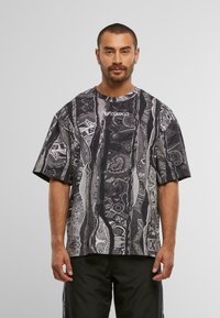 Mand med kort hår og skæg iført en sort og grå mønstret Colucci T-shirt og sorte bukser, stående foran en ensfarvet baggrund.