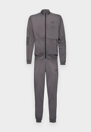 Tuta grigia realizzata in tessuto morbido, caratterizzata da una giacca con zip e colletto alto, con accenti a righe sulle spalle e sui pantaloni.