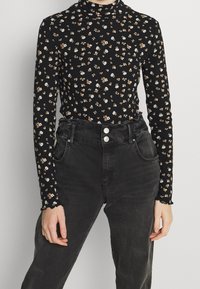 Schwarzes langärmliges Oberteil mit kleinem Blumenmuster in Weiß und Orange, mit hohem Hals und gerippter Textur. Kombiniert mit hochgeschnittenen schwarzen Jeans.