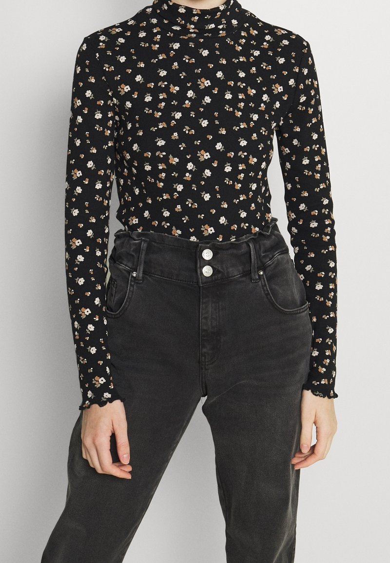 Schwarzes langärmliges Oberteil mit kleinem Blumenmuster in Weiß und Orange, mit hohem Hals und gerippter Textur. Kombiniert mit hochgeschnittenen schwarzen Jeans.
