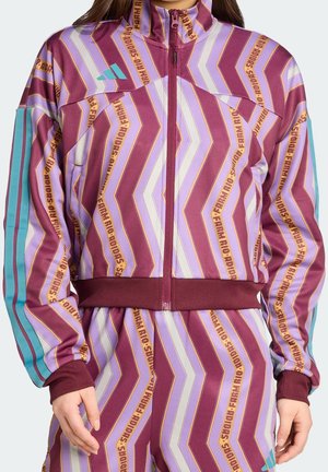 Femme portant un survêtement violet et lavande à motifs avec des bandes bleu sarcelle et des motifs textuels, comprenant une veste zippée et un pantalon assorti.