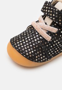 Chaussure en tissu noir avec des motifs d'étoiles métalliques, lacets beiges et semelle en caoutchouc brun clair. Caractérisée par une sangle Velcro pour un attachement facile.