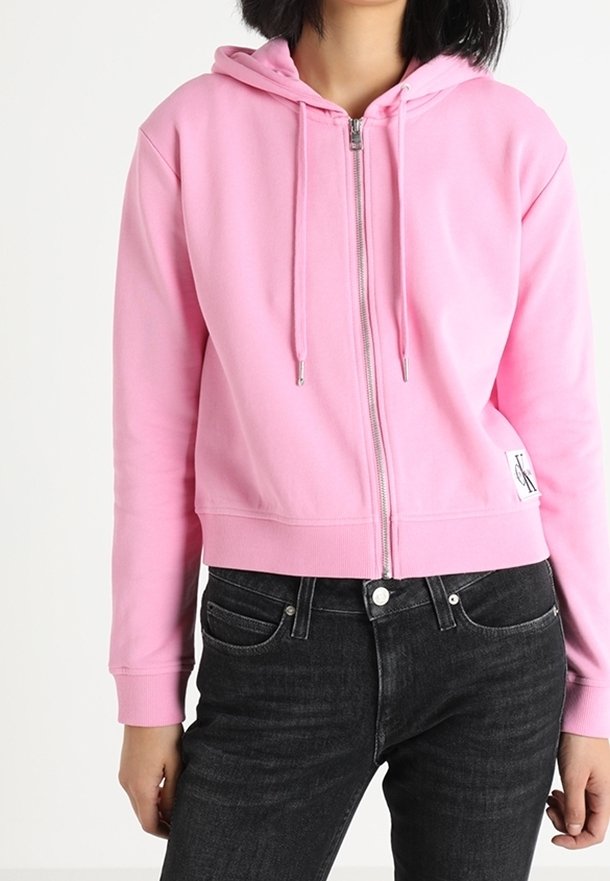 Roze zip-up hoodie gemaakt van zachte stof met een trekkoordcapuchon, cropped ontwerp en een wit logopatch aan de voorkant. Gepaard met donkere jeans.