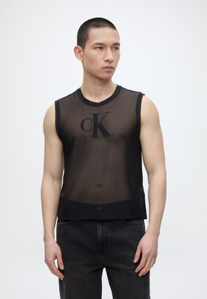 Młody mężczyzna ubrany w czarny, przezroczysty top bez rękawów z logo "cK", w połączeniu z czarnymi dżinsami, stojący na jednolitym białym tle.