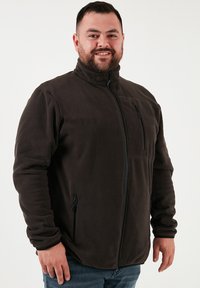 Sötétbarna polárdzseki magas gallérral, elülső cipzárral és két cipzáras oldalzsebbel. Puha tapintás, testhezálló fazon.