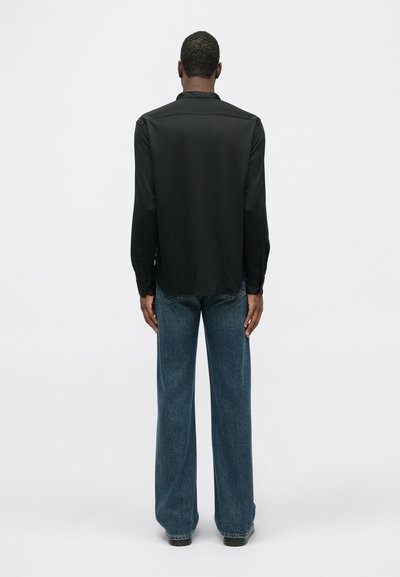 Mies seisoo selin pitkähihainen musta paita ja siniset leveät farkut päällään yksinkertaista valkoista taustaa vasten.