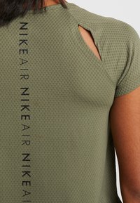 Parte superior de la espalda de una persona que lleva una camiseta Nike de color verde oliva con textura, con un logo vertical negro y un detalle de corte en los hombros.