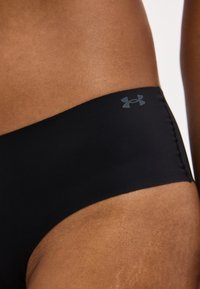 Κοντινό πλάνο του γοφού ενός ατόμου που φοράει μαύρα αθλητικά εσώρουχα Under Armour με λογότυπο στο πλάι, σε απλό φόντο.