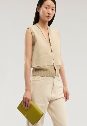 Kvinde iført beige ærmeløs knapvest og bukser, der holder en grøn rektangulær clutch i venstre hånd.