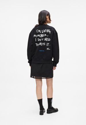 Black oversized sweatshirt with white text on the back. Material: cotton blend. Paired with a black sheer skirt and black footwear.