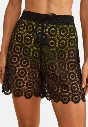 Vrouw draagt zwarte gehaakte shorts met cirkelvormige patronen en een zwarte trekkoord met kwastjes in de taille.