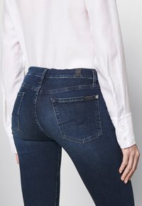 Jeans de mezclilla azul oscuro con un diseño ajustado, que cuentan con dos bolsillos traseros, costuras visibles y una etiqueta cerca de la cintura.