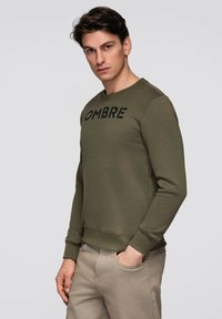 Ciemnozielona bluza z napisem "OMBRE" w kolorze czarnym. Posiada okrągły dekolt, ściągacze na mankietach oraz luźny krój. Noszona z jasnobeżowymi spodniami.