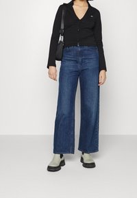 Black long-sleeve button-up shirt, paired with high-waisted wide-leg blue denim jeans and gray ankle boots with black soles. Minimal accessories.