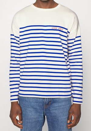 Uomo che indossa una maglietta bianca a maniche lunghe con strisce orizzontali blu e testo rosso "GOOD VIBES", abbinata a jeans blu.