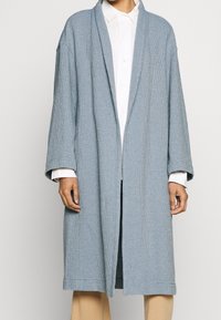 Cappotto tessuto blu chiaro con vestibilità morbida, maniche lunghe, apertura anteriore e superficie texturizzata. Indossato sopra una camicia bianca.