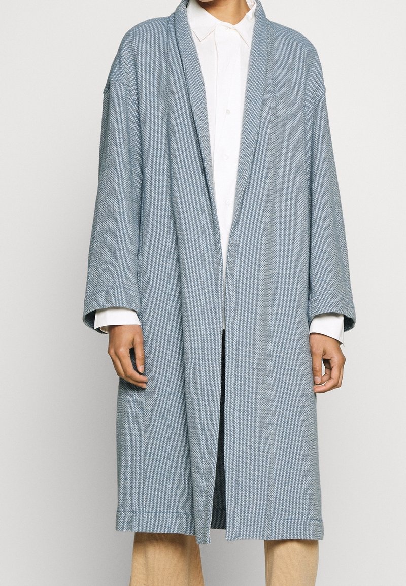 Cappotto tessuto blu chiaro con vestibilità morbida, maniche lunghe, apertura anteriore e superficie texturizzata. Indossato sopra una camicia bianca.
