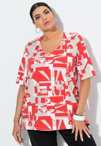 Červené a béžové oversized tričko s výstřihem do V, zdobené geometrickým vzorem. Krátké rukávy a měkká látka, doplněné černými kalhotami.