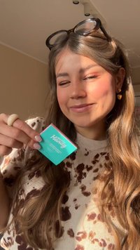 Junge Frau mit langen Haaren und Sonnenbrille auf dem Kopf, lächelnd und eine türkisfarbene Schachtel mit der Aufschrift "Hello Handy" in einem sonnenbeschienenen Raum haltend.