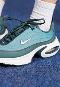 Tökéletesen összehangolt türkiz és sötétzöld Nike sportcipők, elegáns kialakítással, fehér talppal, látható légpárnázással és fűzőkkel. Texturált anyag ezüst kiegészítőkkel.