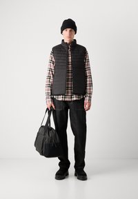 Um colete acolchoado preto sobre uma camisa xadrez, combinado com jeans escuros. O modelo segura uma bolsa preta; usa um gorro preto e sapatos grossos pretos.