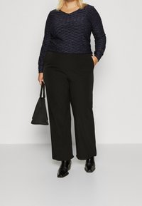 Mörkblå långärmad topp med V-krage och strukturerat mönster; tillsammans med svarta vida byxor och svarta ankelstövlar; håller en liten svart handväska.