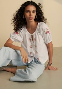 Hvid kortærmet bluse med broderede blomster mønstre og knaplukning. Matchet med lyseblå brede jeans. Glat stofstruktur.