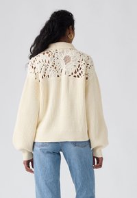 Pull en tricot couleur crème avec un panneau arrière en dentelle crochetée, une texture côtelée et des manches ballon. Porté avec un jean bleu clair.