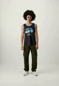 Jovem com cabelo cacheado grande, usando óculos de sol brancos, uma t-shirt gráfica preta sem mangas, calças cargo olivas e ténis brancos, em pé.