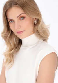 Blond kvinde med blå øjne iført gyldne øreringe og en ærmeløs hvid turtleneck, der smiler blidt mod en hvid baggrund.