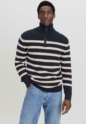 Gebreide trui met een hoge kraag, voorzien van marineblauwe en crèmekleurige horizontale strepen. Ribbelstructuur en een halve rits. Gedragen met lichtblauwe jeans.