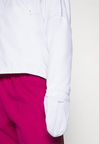 Personne portant une veste blanche avec une moufle blanche intégrée avec un petit logo Nike, associée à un pantalon magenta vif.