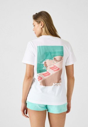 Witte katoenen t-shirt met een grote, kleurrijke strandscène op de achterkant, met roze ligstoelen, blauw water en lichtbruine stenen.