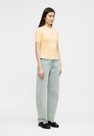 Γυναίκα που στέκεται, φορώντας ένα πλεκτό κοντομάνικο μπλουζάκι ωχρού κίτρινου χρώματος με γιακά, ανοιχτό μπλε τζιν με φαρδιές πιέτες και μαύρα μοκασίνια.