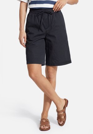 Persoon draagt marineblauwe shorts met trekkoord, wit en marineblauw gestreept topje, staand met gekruiste benen en bruine sandalen met gesp.