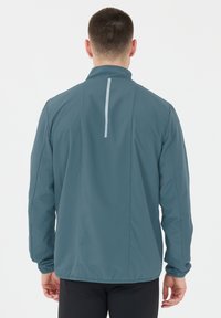 Lättvikts, teal färgad vindjacka med hög krage, långa ärmar och en reflexvertikal rand på ryggen. Slät tygkvalitet.