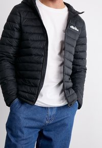 Czarna kurtka puchowa z kapturem, z pikowaną teksturą, zamkiem błyskawicznym i małym logo. Nosi się ją na białą koszulę i niebieskie dżinsy.