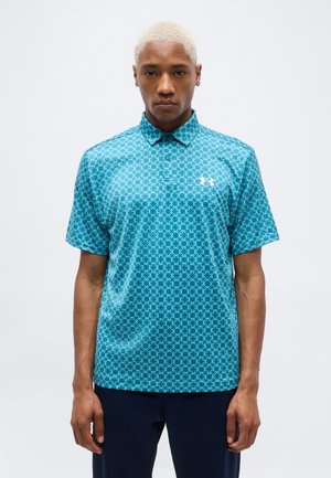 Muž s krátkymi blond vlasmi, oblečený v priliehavej tyrkysovej polokošeli s vzorom a tmavých nohaviciach, stojaci pred jednofarebným svetlým pozadím.