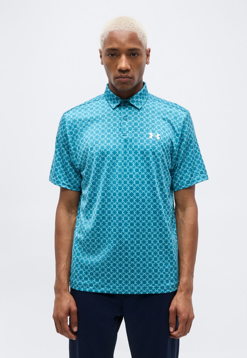 Άνδρας με κοντά ξανθά μαλλιά, ντυμένος με εφαρμοστό τυρκουάζ πόλο με σχέδια και σκούρο παντελόνι, στέκεται μπροστά σε απλό ανοιχτό φόντο.