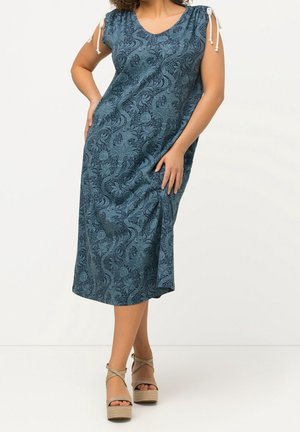 Vrouw draagt een blauwe mouwloze midi-jurk met bloemenpatroon, gestrikte schouderbandjes en beige plateau sleehaksandalen, staand tegen een witte achtergrond.