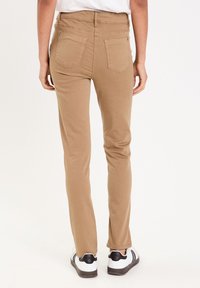 Pantalon beige slim-fit avec une taille mi-haute, deux poches arrière et une texture en mélange de coton lisse, associé à des baskets noires et blanches.