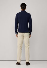 Man staat tegenover een muur en draagt een marineblauwe lange mouwen polo, beige broek en donkerbruine schoenen op een lichtgrijze achtergrond.