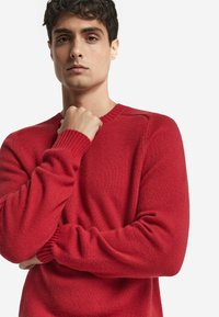 Röd stickad tröja med rund hals och ribbade ärmslut. Har en mjuk textur och figurnära design som framhäver en tight passform på armarna.