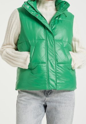 Personne portant une doudoune verte brillante par-dessus un pull côtelé blanc et un jean bleu clair, les mains dans les poches de la doudoune.