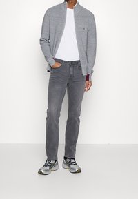 Cárdigan gris con cremallera sobre una camiseta blanca, combinado con jeans grises y zapatos deportivos plateados con detalles amarillos. Diseño minimalista y casual.