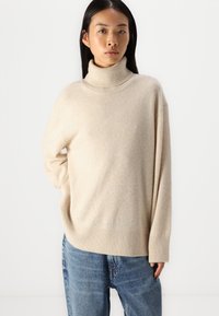 Beige turtleneck sweater in soft knit material, featuring a relaxed fit, ribbed hem, and cuffs, paired with light blue denim jeans.