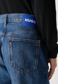 Sinine denimhari, millel on oranž õmblus, ruudukujuline tagatasku ja sinine logotäht "HUGO" vöökoht. 