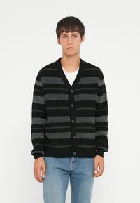 Black and grey striped cardigan with a v-neck, button closure, and ribbed cuffs. Worn over a white t-shirt and paired with blue jeans.